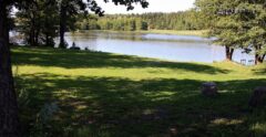 Bilde av en stor plen med trær på hver side og vannet nedenfor.  Bakenfor vannet, på andre siden vises en åker og masse trær. Viser Rødsund badeplass.